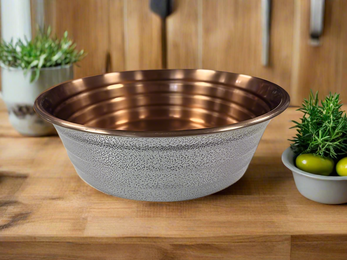 A white ribbed dog bowl made of copper, placed on a wooden surface with a plant in the background.