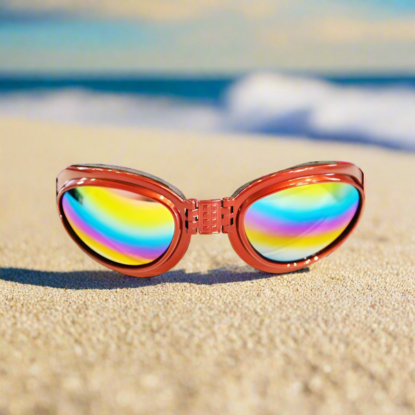 A pair of Doggles with multicolored lenses and red frame lying on a sandy beach with a blurred background.