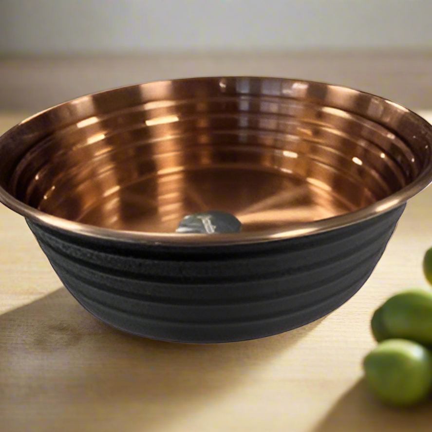 A ribbed black dog bowl with a copper interior, placed on a wooden surface next to some green olives.