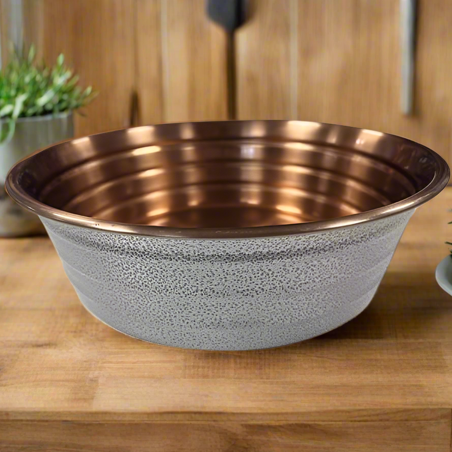A white ribbed dog bowl made of copper, placed on a wooden surface with a plant in the background.
